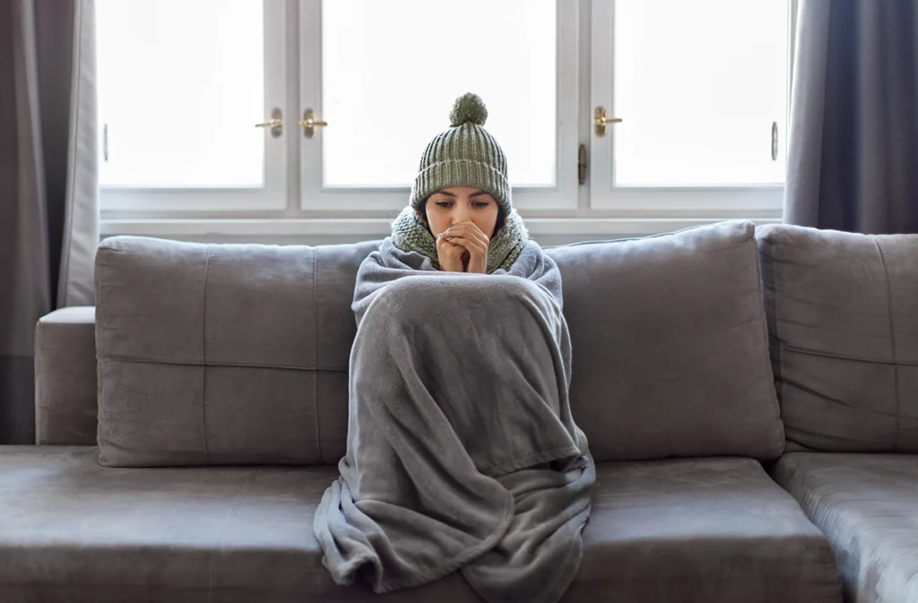 Woman sitting on a couch wrapped in a blanket and winter hat while appearing cold indoors. Best for blogs about no heat, heating problems, furnace issues, and winter indoor discomfort.