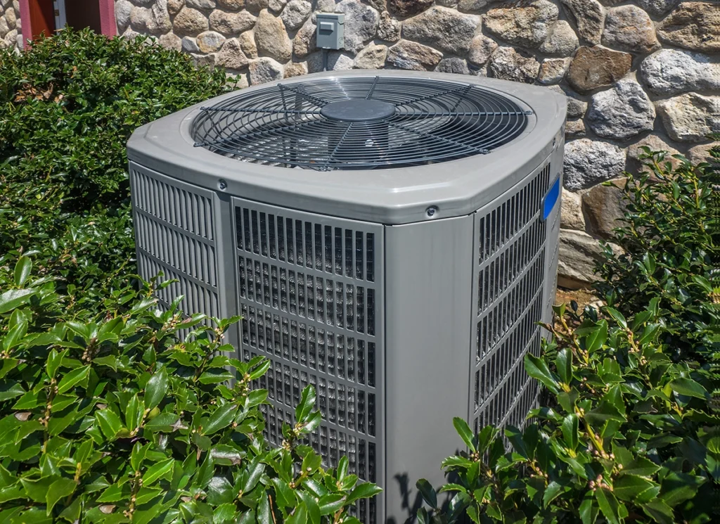 Outdoor residential air conditioning condenser unit surrounded by landscaping near a stone wall.
