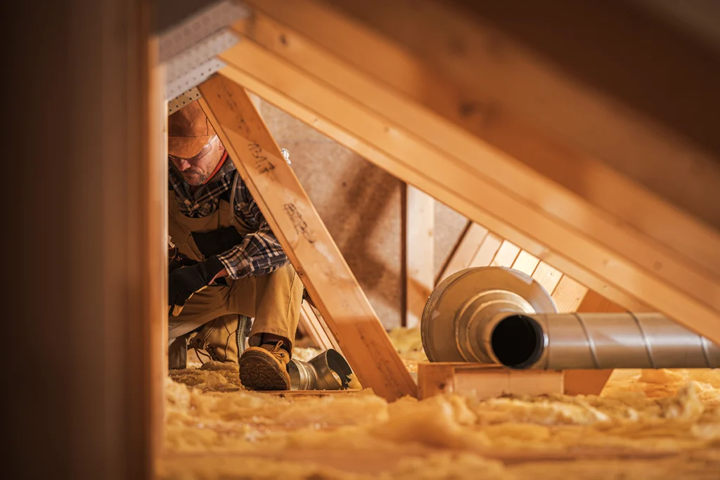 Technician working in an attic near exposed ductwork and insulation in a residential space. Best for blogs about attic ductwork, duct repair, HVAC installation, insulation, and airflow problems.